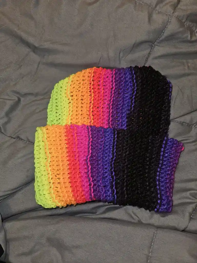 A hand-crocheted, rainbow-striped beanie with vertical stripes of lime green, orange, red, pink, purple, and black, folded…