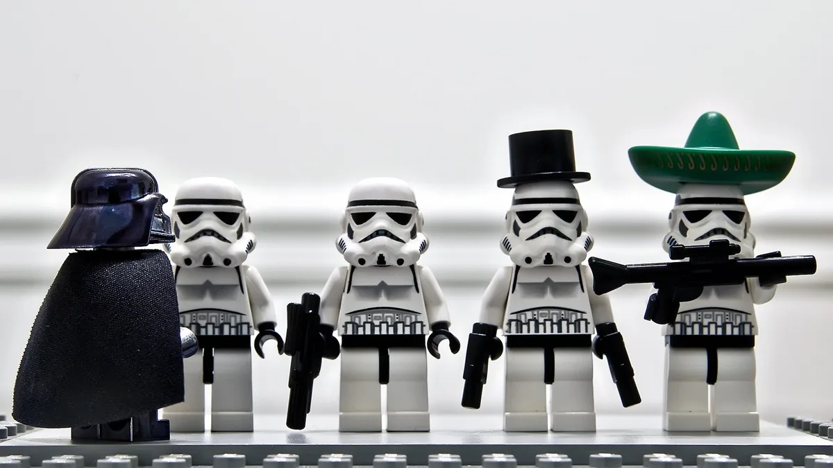 A Lego Darth Vader stands facing four Lego Stormtroopers. Three of the Stormtroopers wear standard helmets, while the fourth…