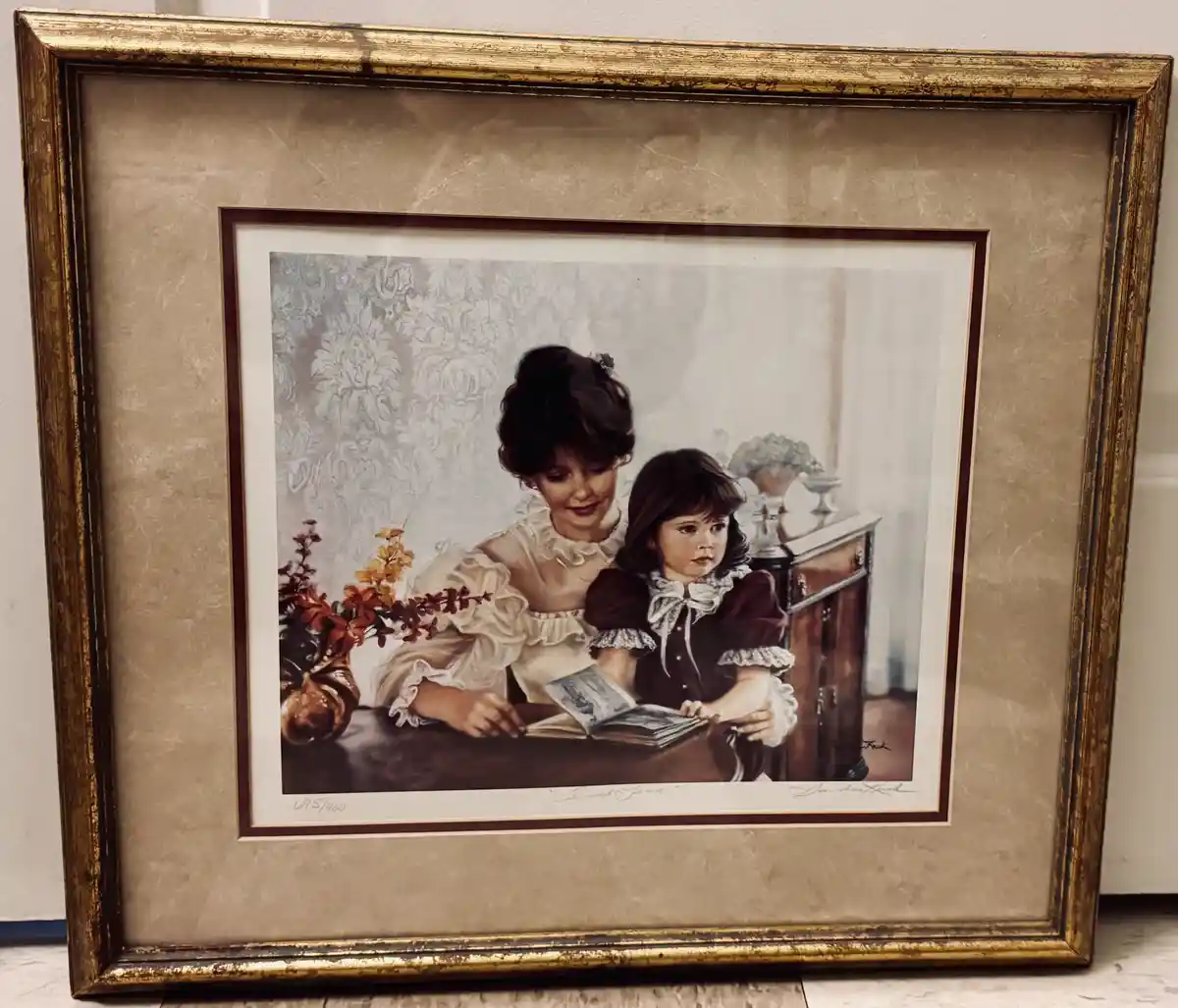 A framed vintage-style print depicts a woman in a white ruffled dress reading a book to a young girl in a dark dress with…