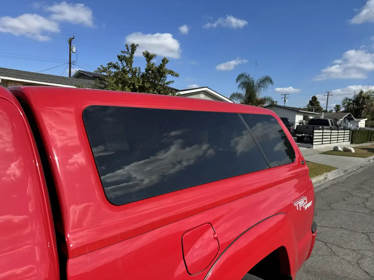 A close-up photo of the rear of a shiny red pickup truck, focusing on its tinted rear window which reflects the blue sky and…