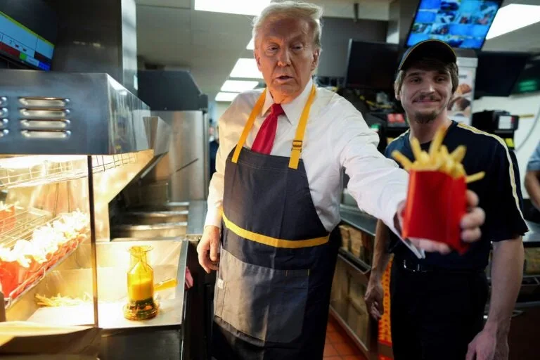 A man resembling Donald Trump, wearing a white shirt, red tie, and a dark apron with yellow straps, stands in a fast-food…