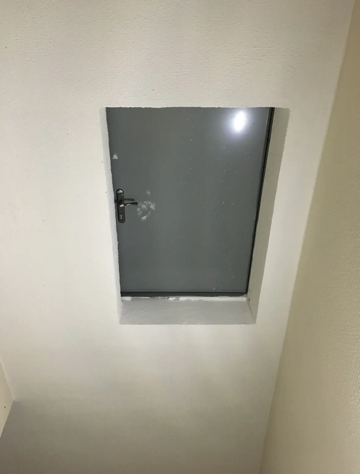A small, grey metal door set into a white wall, appearing unusually tiny and embedded flush with the surrounding plaster…