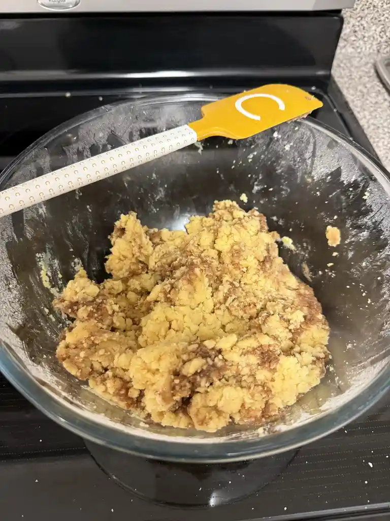 A clear glass mixing bowl sits on a black stovetop, containing a lumpy, crumbly, yellowish-brown mixture that looks like…