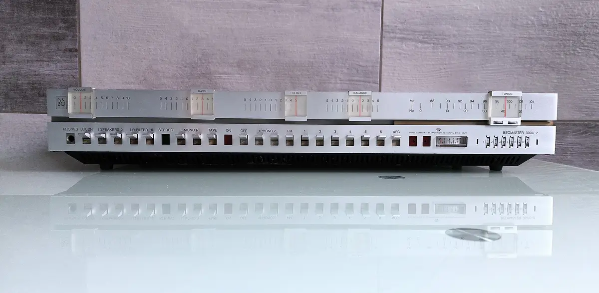 A silver Bang & Olufsen Beomaster 3000-2 vintage stereo receiver sits on a glass surface, its numerous knobs and switches…