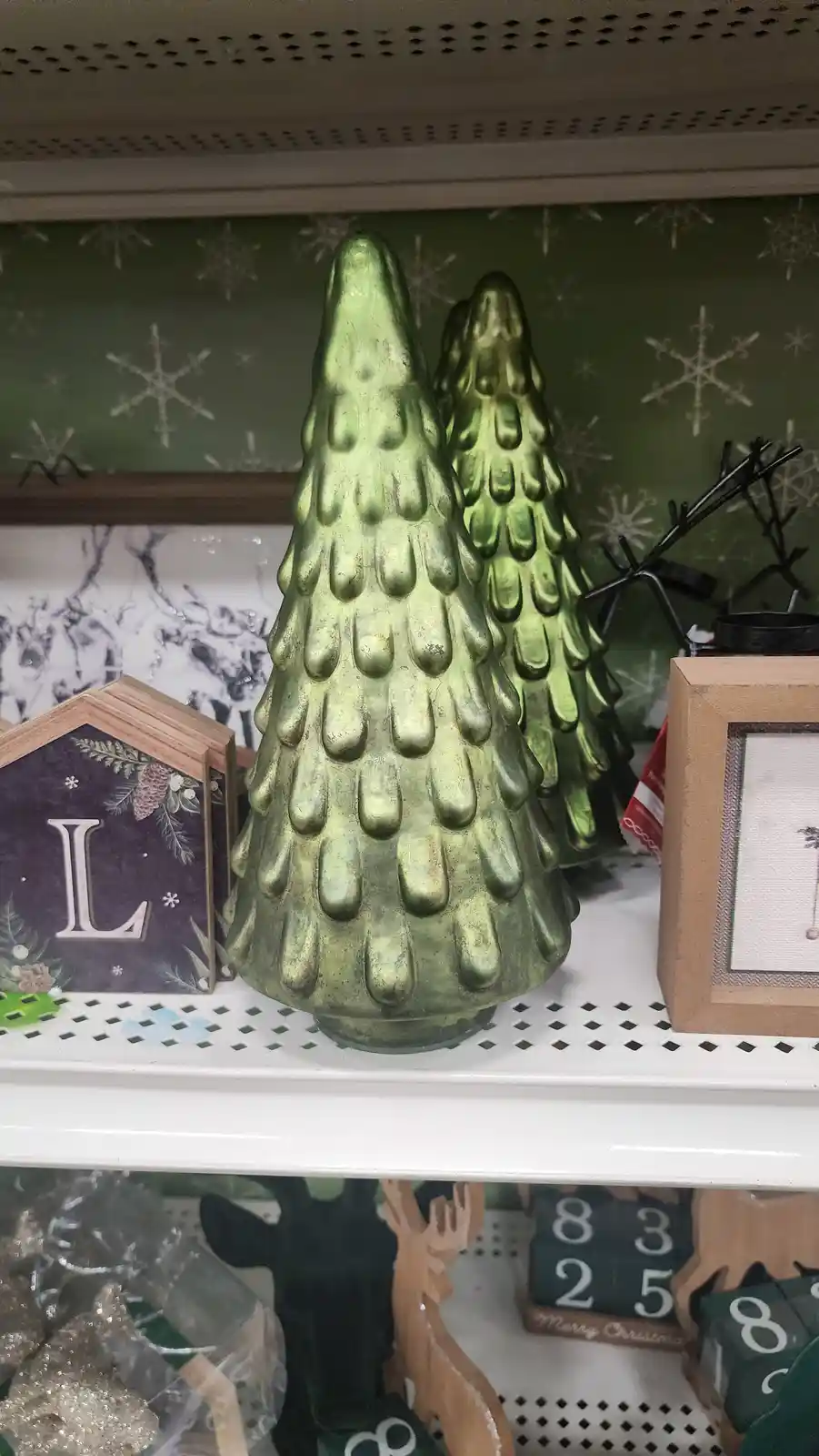 A close-up photo of two green, textured, cone-shaped Christmas tree decorations on a store shelf, surrounded by other…