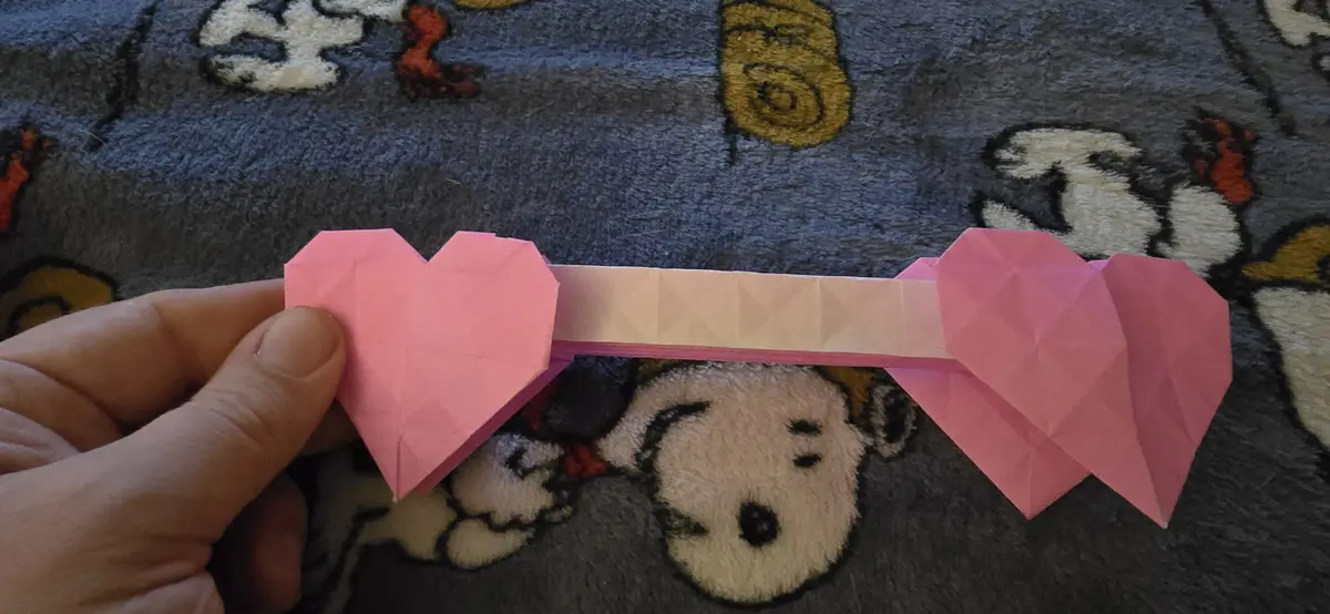 A hand holds a pink origami object shaped like a sword, with heart-shaped blades at both ends and a light pink handle…