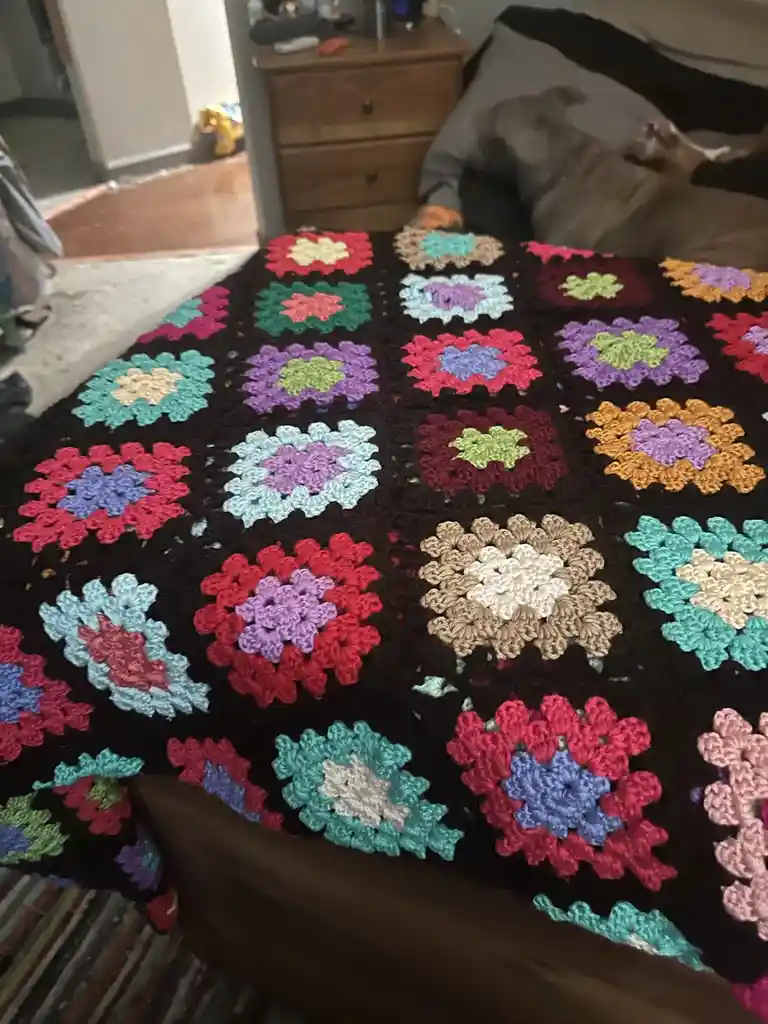 A colorful, hand-crocheted granny square blanket with multicolored floral patterns covers a bed. In the background, a dog…