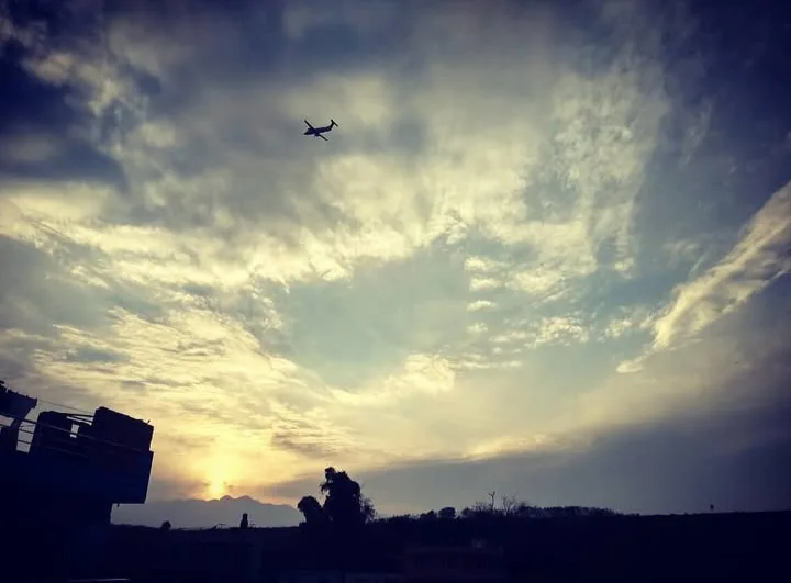 A silhouette of a small airplane flying against a dramatic sunset sky with scattered clouds. Below, dark silhouettes of…