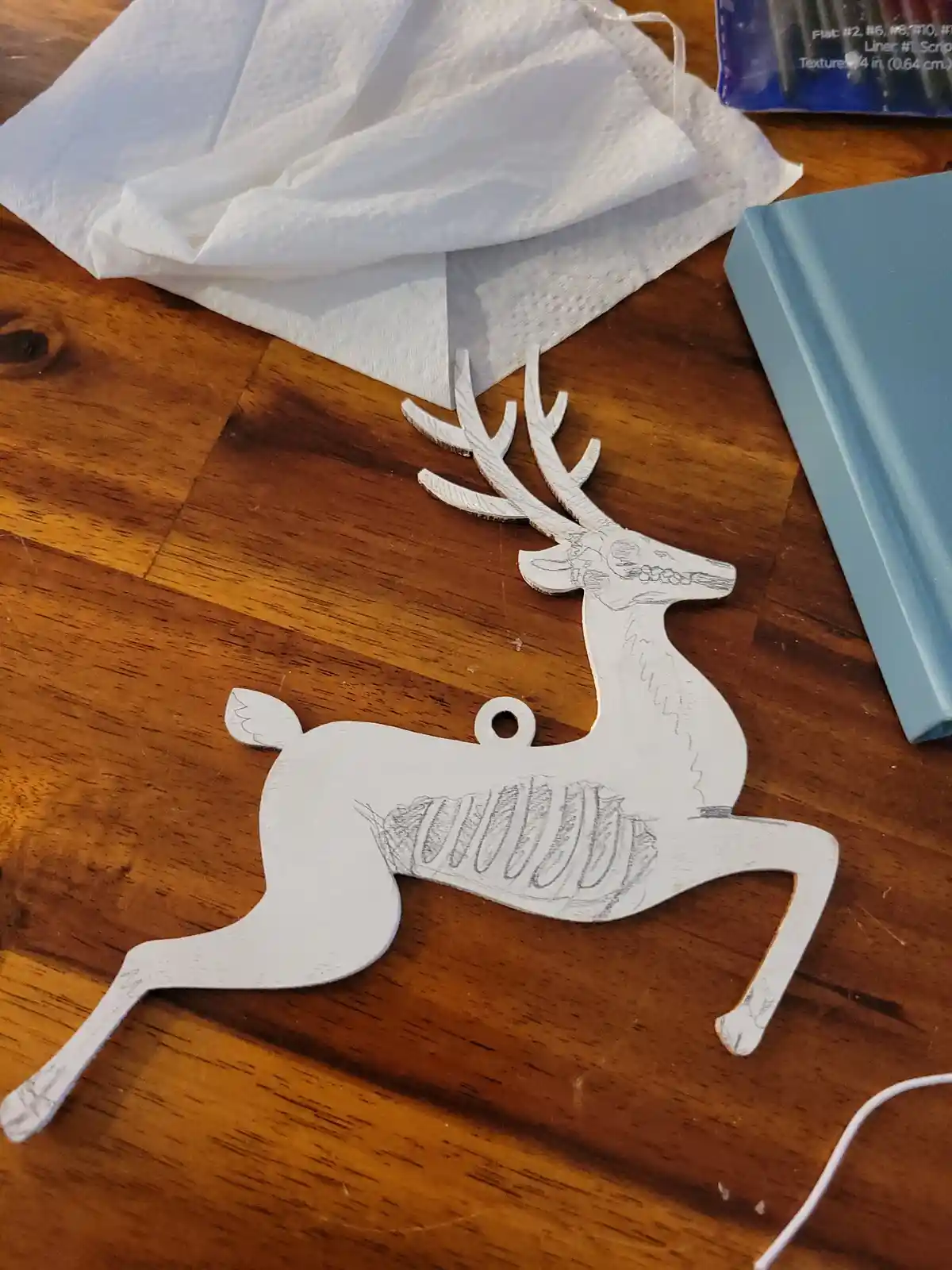 A white-painted wooden reindeer ornament lies on a wooden table, with pencil sketches of a ribcage and skull drawn on its…