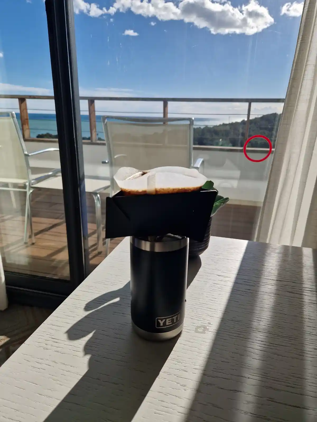 A black YETI tumbler with a pour-over coffee setup sits on a white table indoors, with a bright view of a balcony, ocean…