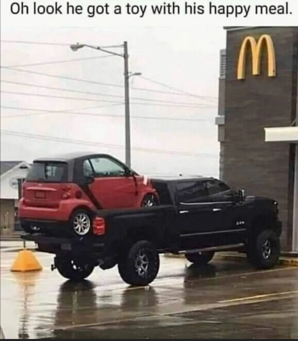 A red Smart car is visibly loaded into the bed of a large black pickup truck parked in front of a McDonald's restaurant. The…