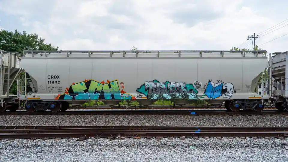 A white freight train car covered in colorful graffiti sits on railway tracks under a cloudy sky. The graffiti includes…
