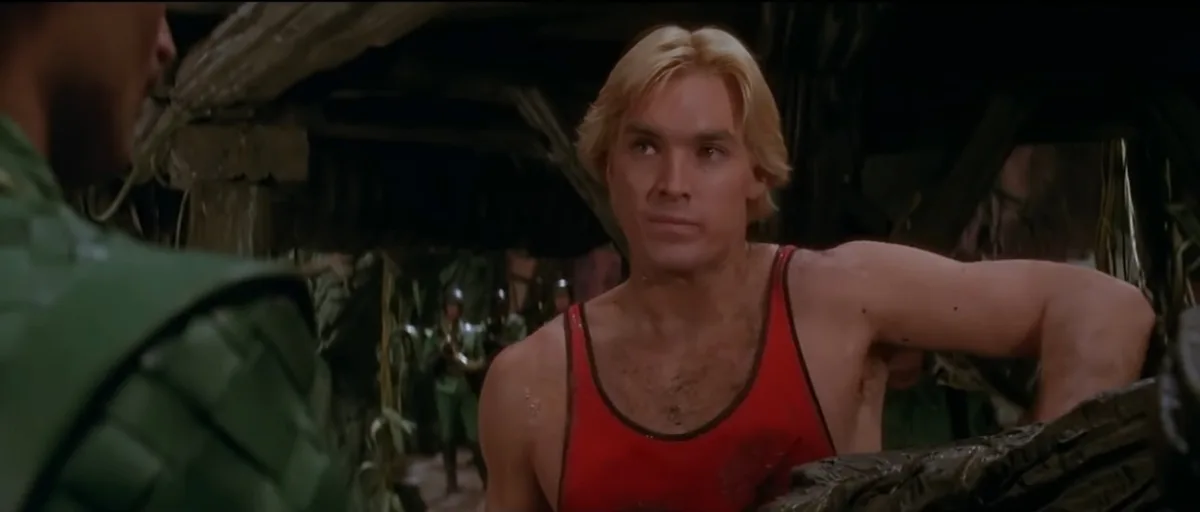 a man with blonde hair and a red tank top looks at someone off-screen with a confused, slightly disgusted expression. He is…