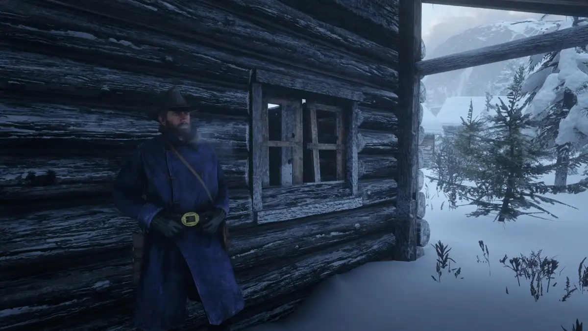 A man in a blue winter coat and hat stands outside a snow-covered log cabin in a snowy, mountainous landscape. He has a…