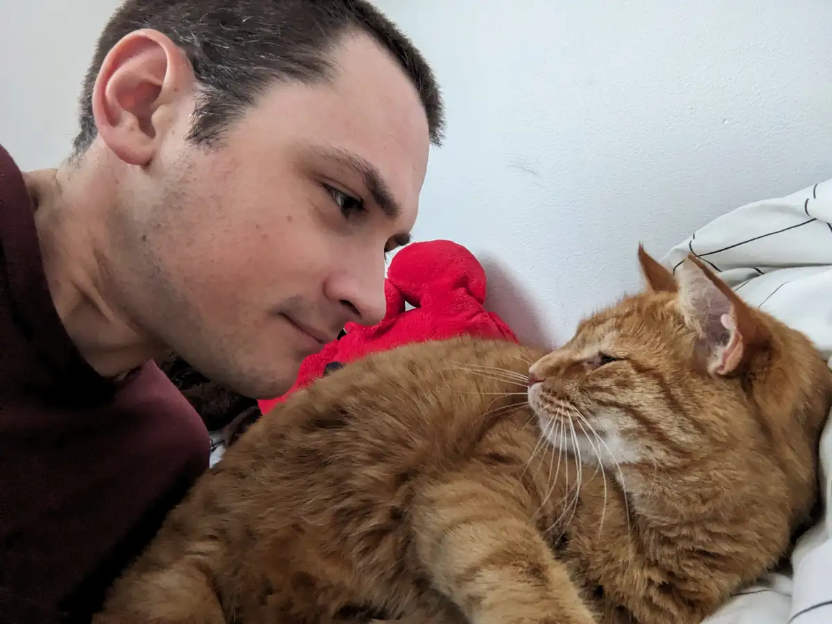 A man in a maroon shirt lies on a bed next to a large orange tabby cat, both looking at each other with affectionate…