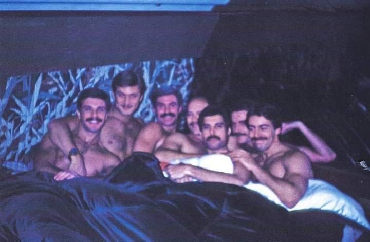 A grainy, low-light photo shows seven shirtless men with mustaches lounging together on a bed under a dark blanket. They are…