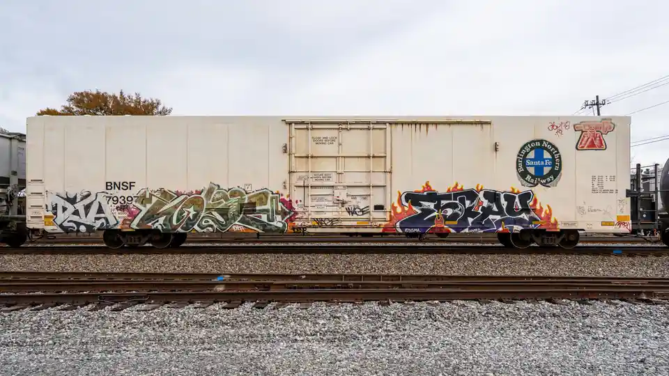 A white Burlington Northern Santa Fe freight train car sits on tracks, covered in colorful graffiti tags. The car has…