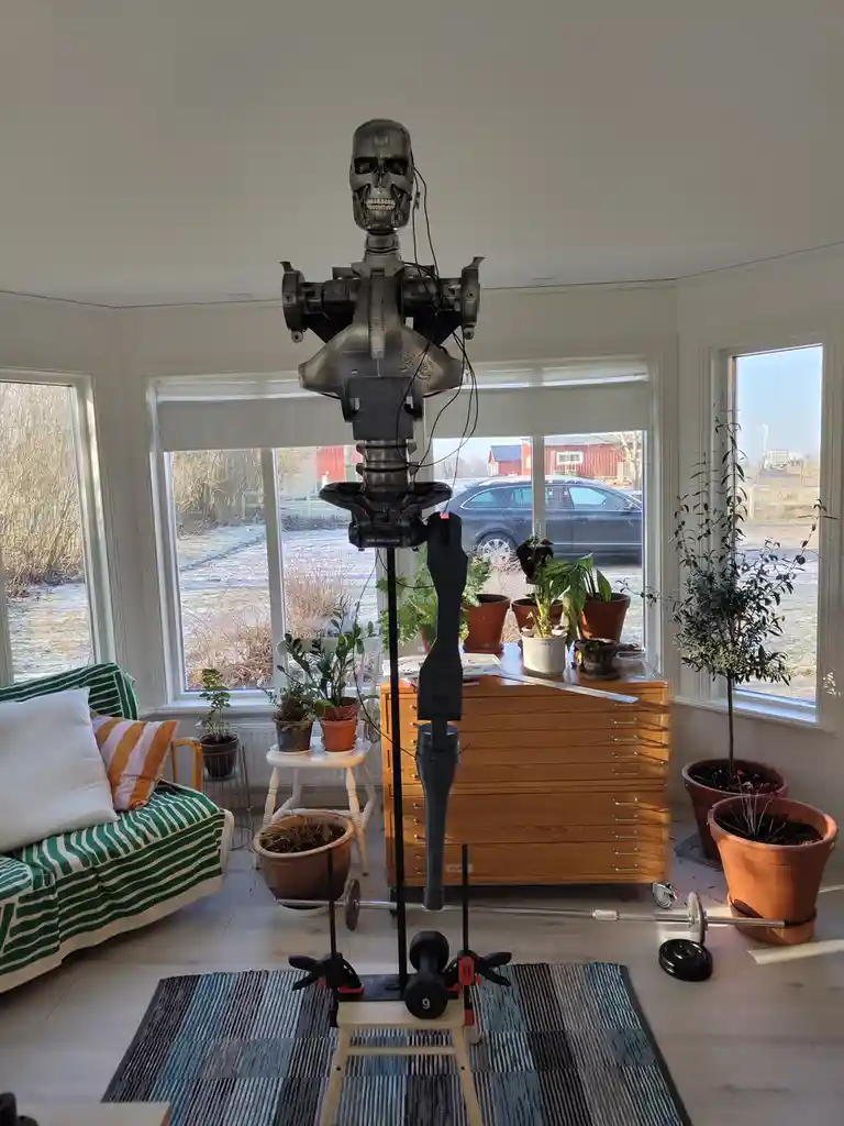 A Terminator-style robot head and torso are mounted on a tall black pole in a bright, cozy living room filled with potted…