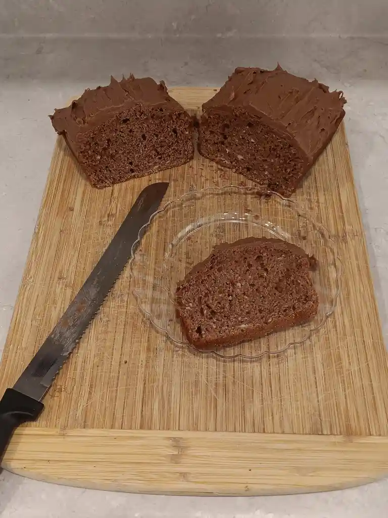A wooden cutting board holds two large, frosted chocolate cake slices and one smaller slice on a clear glass plate, with a…