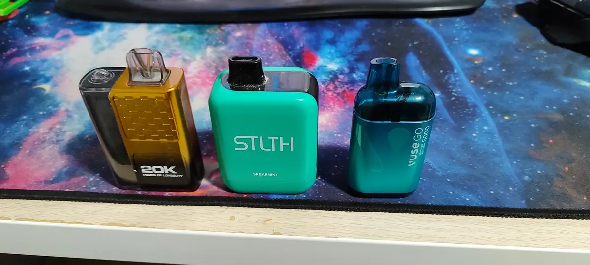 Three different vape pens are lined up on a colorful galaxy-patterned mousepad: a gold and black 20K, a teal STLTH…