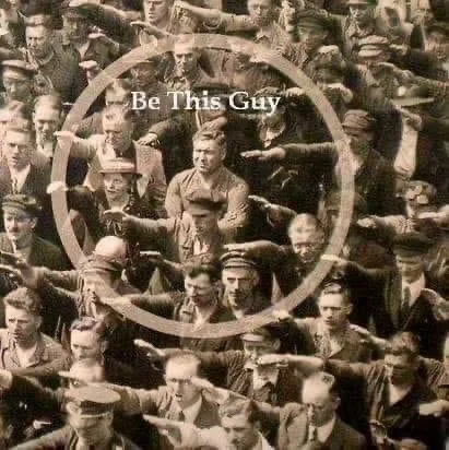 A black-and-white historical photograph shows a dense crowd of uniformed men, many saluting with their right arms extended…