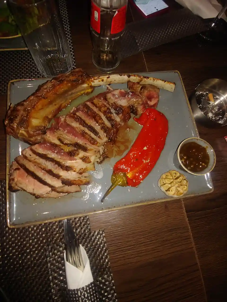 A plated meal featuring a large, sliced grilled ribeye steak with a bone, accompanied by a roasted red pepper, roasted…