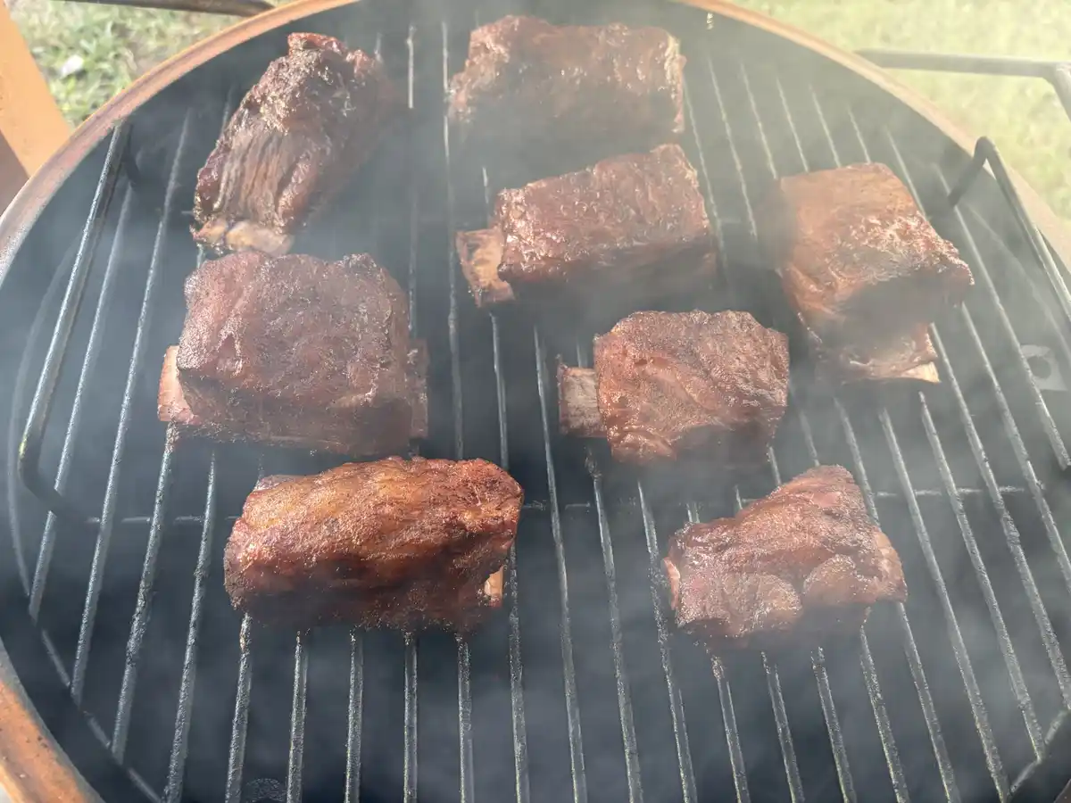 A close-up photo shows several pieces of meat, likely ribs, sizzling on a black metal grill grate over a smoker, with…