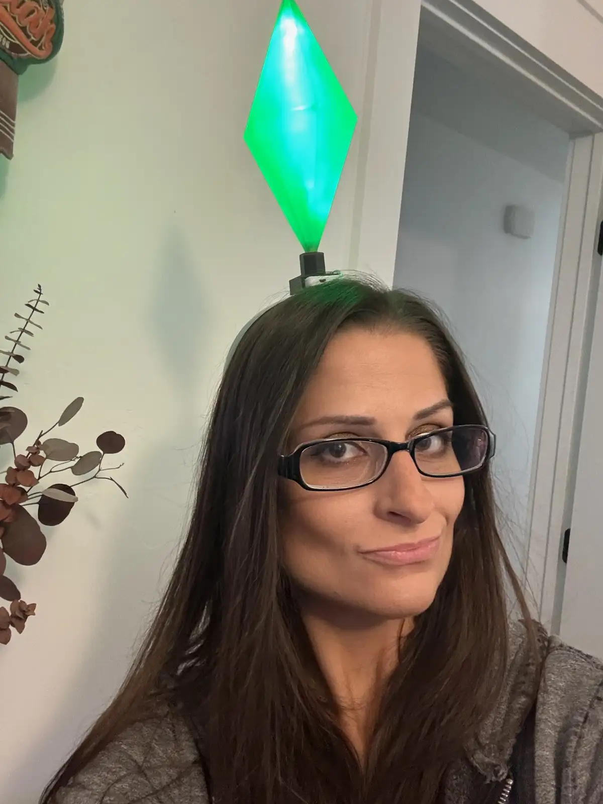A woman with dark hair and glasses looks at the camera with a slight smirk while wearing a glowing green diamond-shaped…