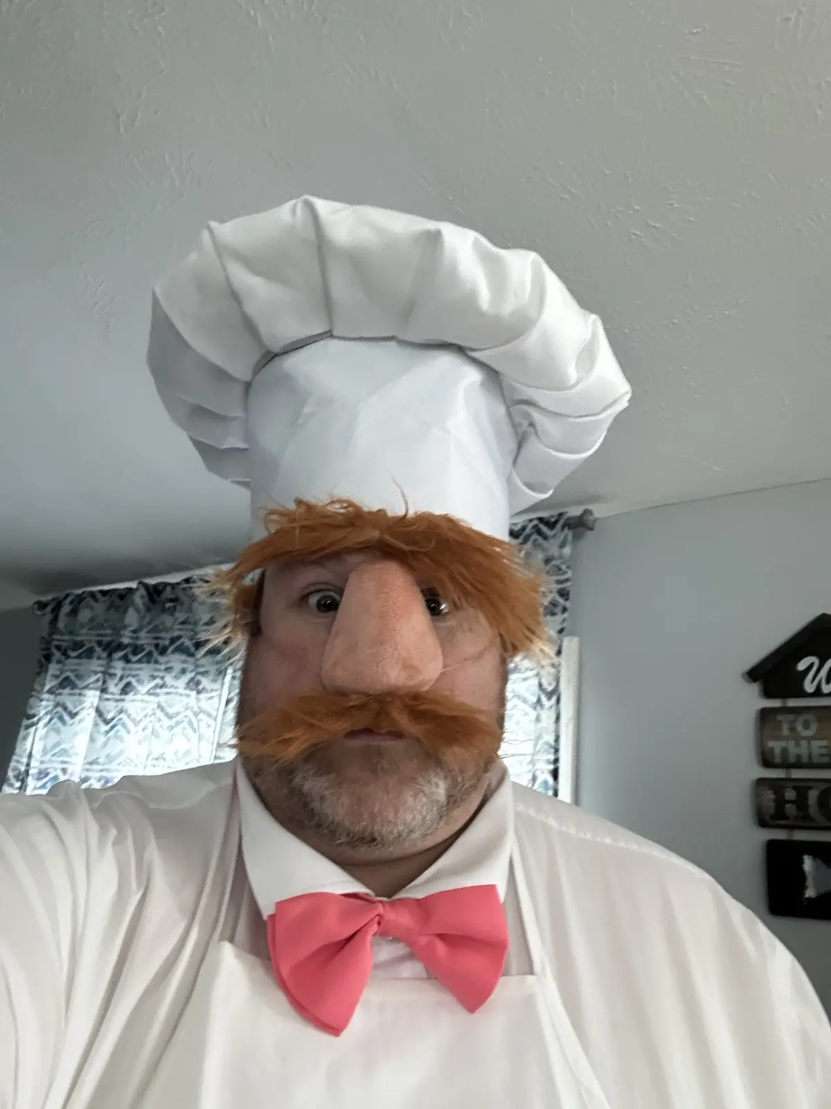 A person wearing a large white chef's hat, a red bow tie, and a white apron, with a large, exaggerated puppet head featuring…
