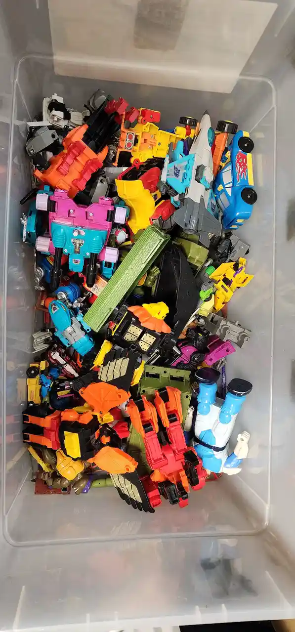 A top-down view of a clear plastic bin overflowing with a chaotic pile of colorful, broken, and disassembled Transformers…