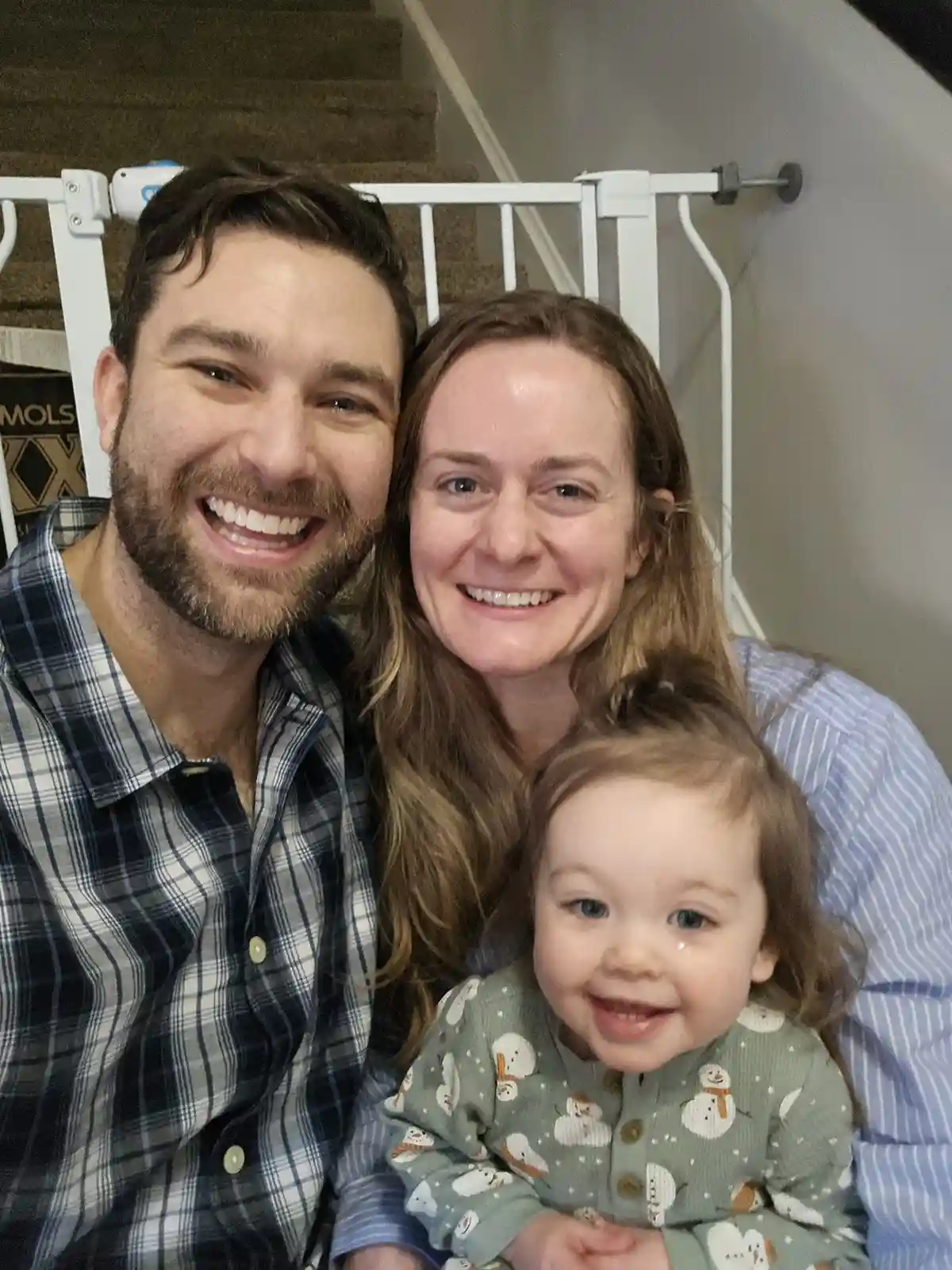 A family photo of a smiling man, woman, and toddler in front of a white baby gate on stairs. The toddler has a tear on their…