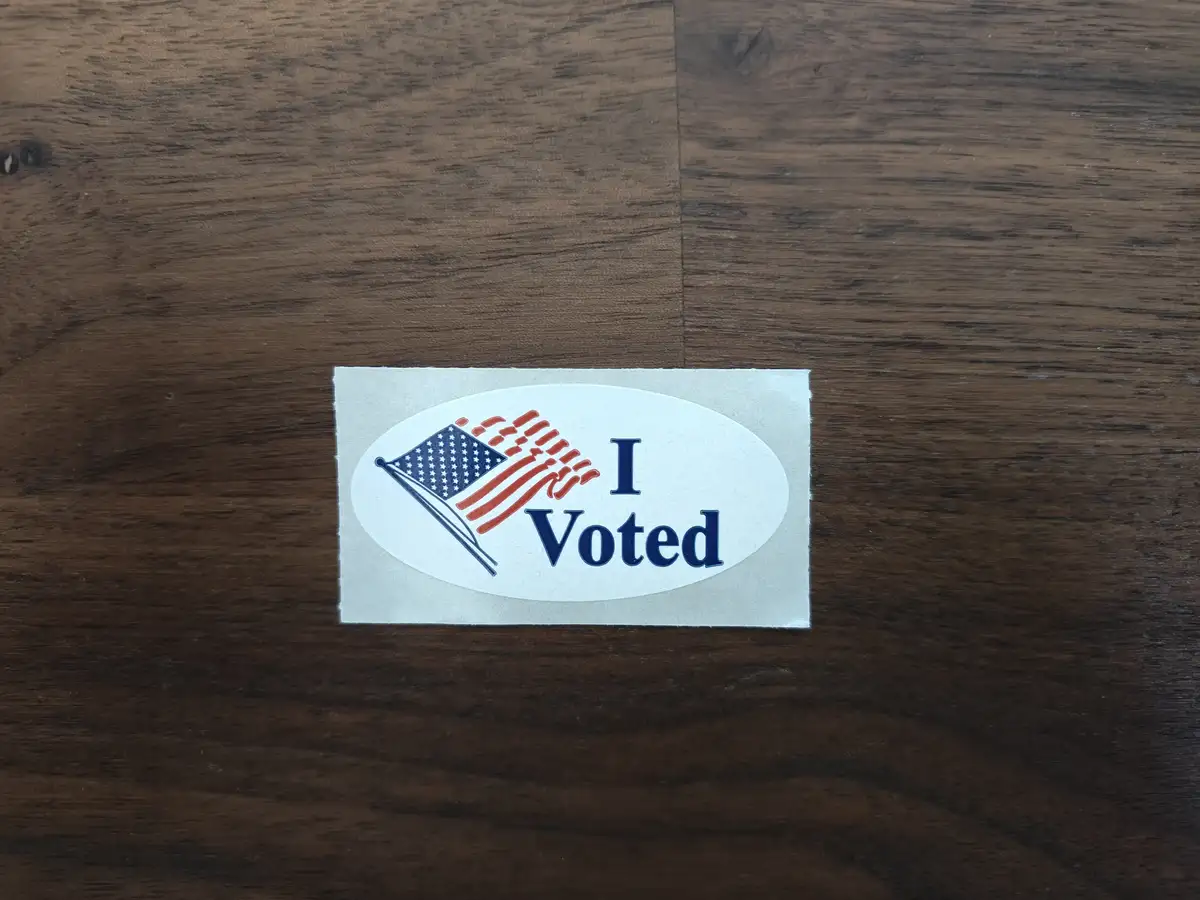 A standard American 'I Voted' sticker featuring a small American flag and the text 'I Voted' in blue, placed on a dark…