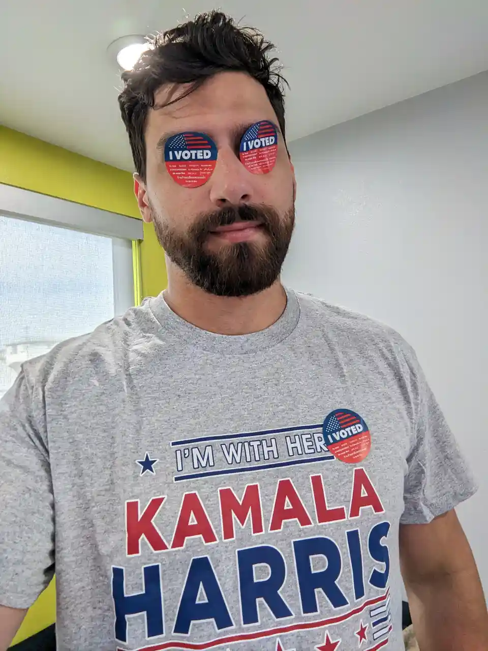 A man with a beard wears a gray t-shirt that says 'I'M WITH HER KAMALA HARRIS' and has an 'I VOTED' sticker on his chest. He…