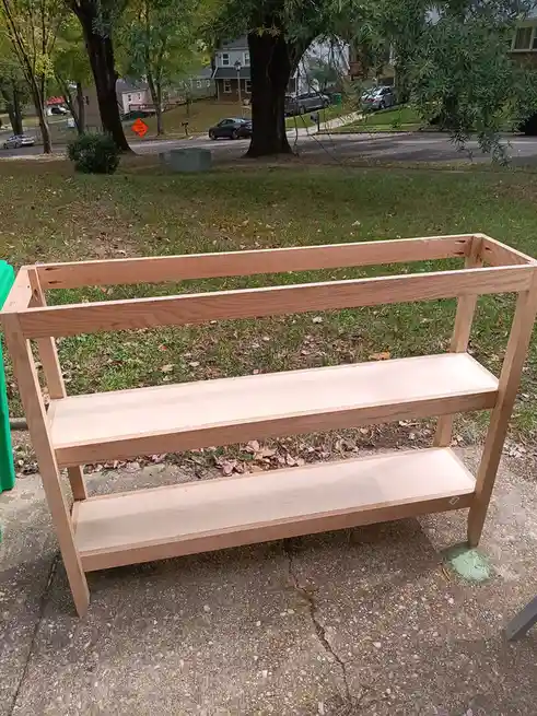 A freshly built, unfinished wooden three-tiered shelf sits outdoors on a concrete driveway, with a grassy yard and suburban…