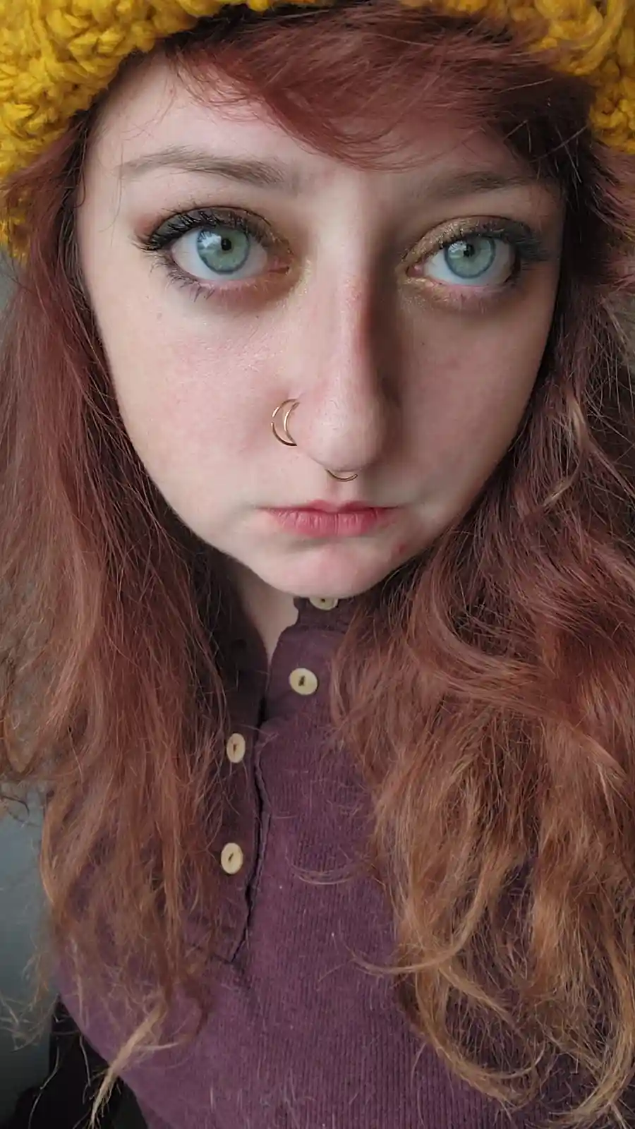 A close-up selfie of a person with reddish-brown hair, wearing a yellow knitted headband and a purple button-up shirt. They…