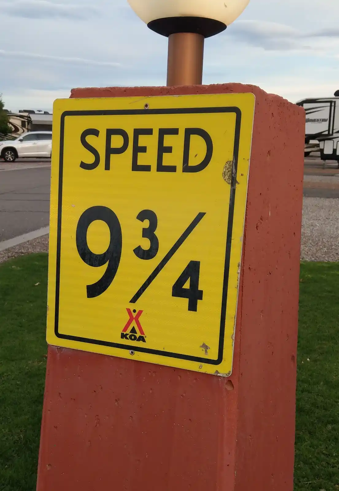 A yellow rectangular speed limit sign mounted on a red post shows the text 'SPEED 9 3/4' in bold black letters, with the KOA…