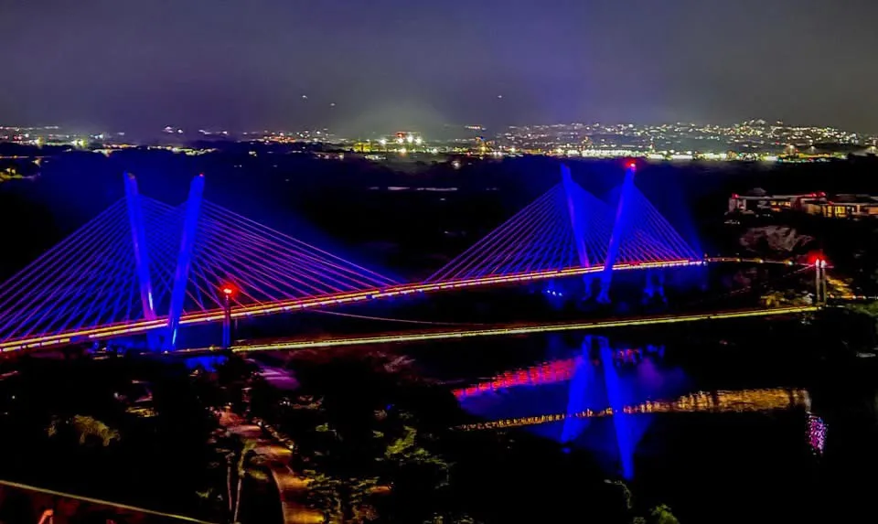 A nighttime photo of a modern cable-stayed bridge illuminated with blue and red lights, spanning a dark river with city…