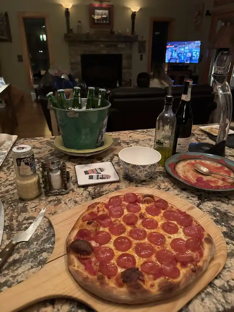 A large pepperoni pizza sits on a wooden board on a granite countertop, surrounded by beer bottles in a bucket, wine, and a…