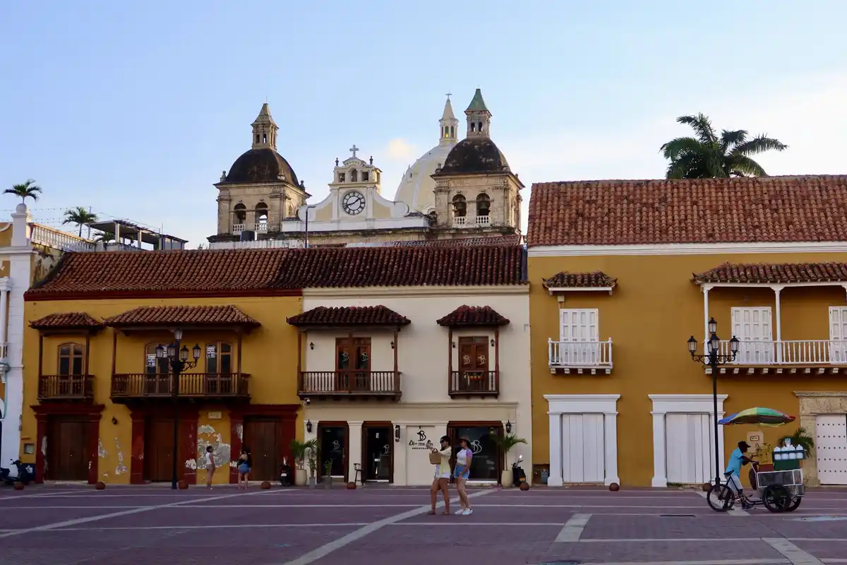A scenic photo of a colonial plaza with yellow buildings, red-tiled roofs, and a church with domes and a clock tower under a…
