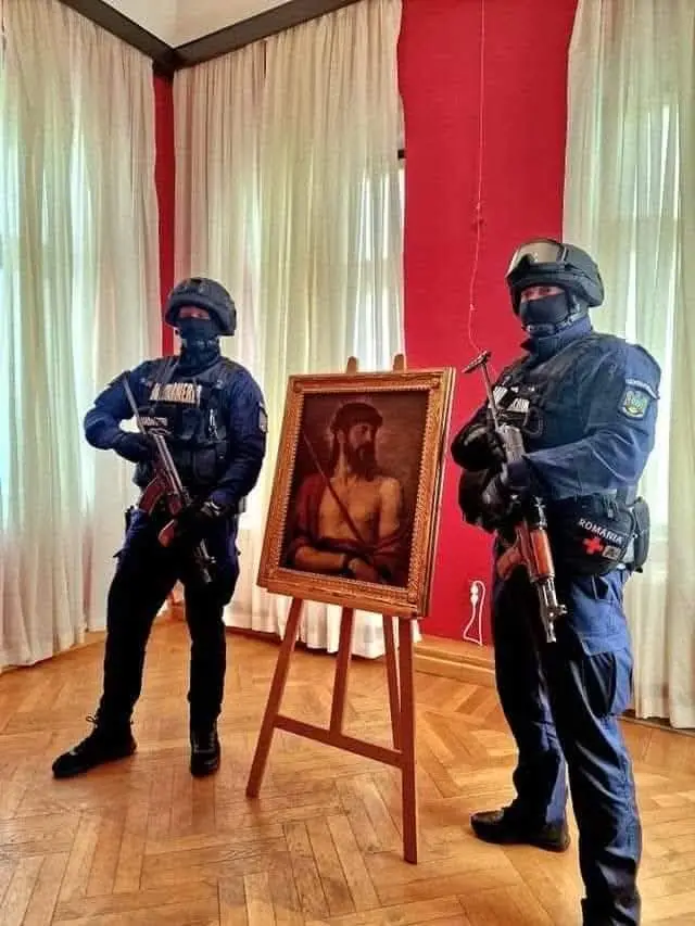 Two heavily armed Romanian police officers in tactical gear stand on either side of a wooden easel displaying a framed…