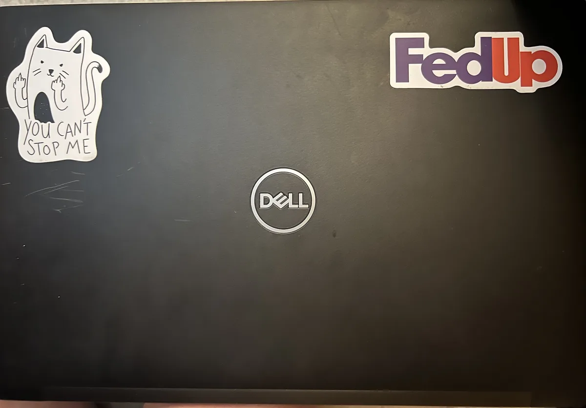 A black Dell laptop lid shows two stickers: a white cartoon cat with raised middle fingers and the text 'YOU CAN'T STOP ME'…