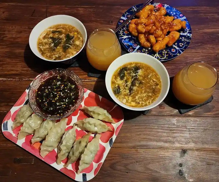 An overhead shot of a wooden table with two bowls of egg drop soup, a plate of sesame chicken, a plate of dumplings with…