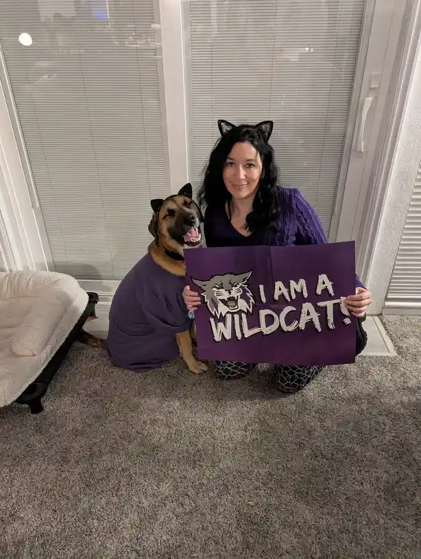 A woman and a dog, both wearing black cat ears and matching purple shirts, kneel on a carpeted floor in front of a window…