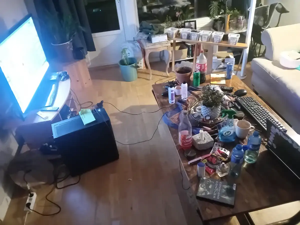 A messy living room with a cluttered coffee table full of drinks, cleaning supplies, gaming gear, and a 'Command & Conquer'…