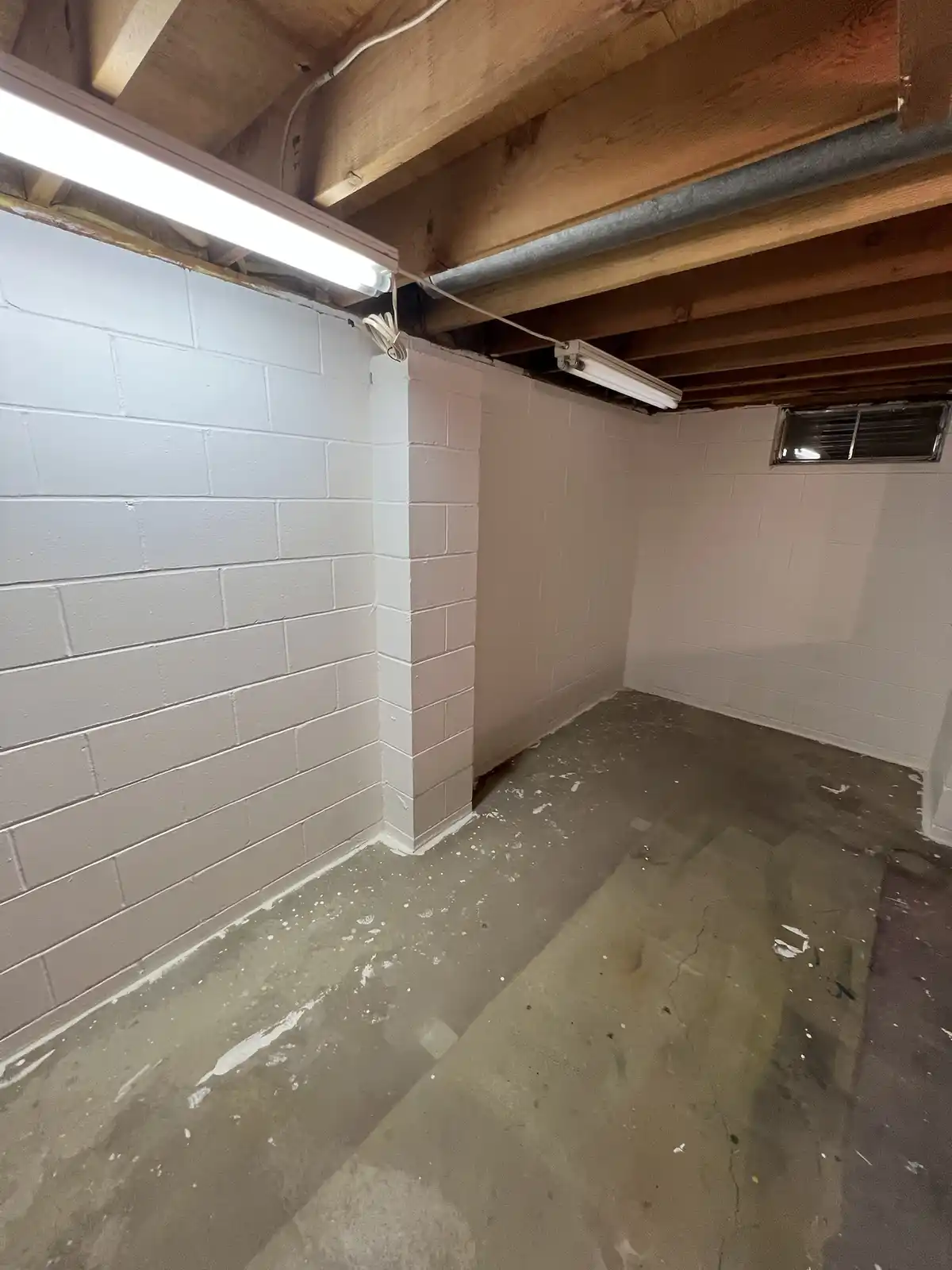 An empty, unfinished basement with white-painted cinder block walls, exposed wooden beams and pipes on the ceiling, and a…
