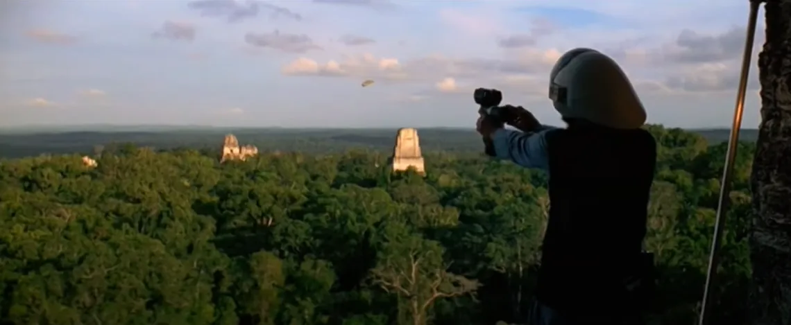 A person wearing a white helmet and dark vest stands on a high vantage point overlooking a dense jungle with ancient stone…