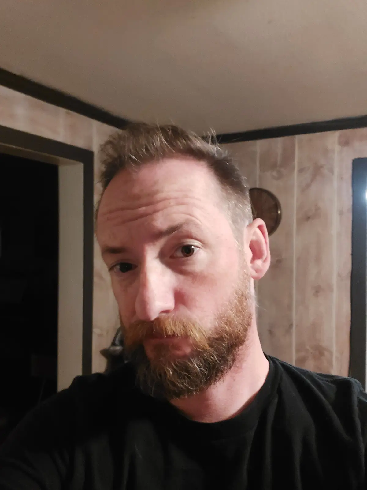 A man with a reddish-brown beard and short hair looks directly at the camera with a skeptical expression. He is wearing a…