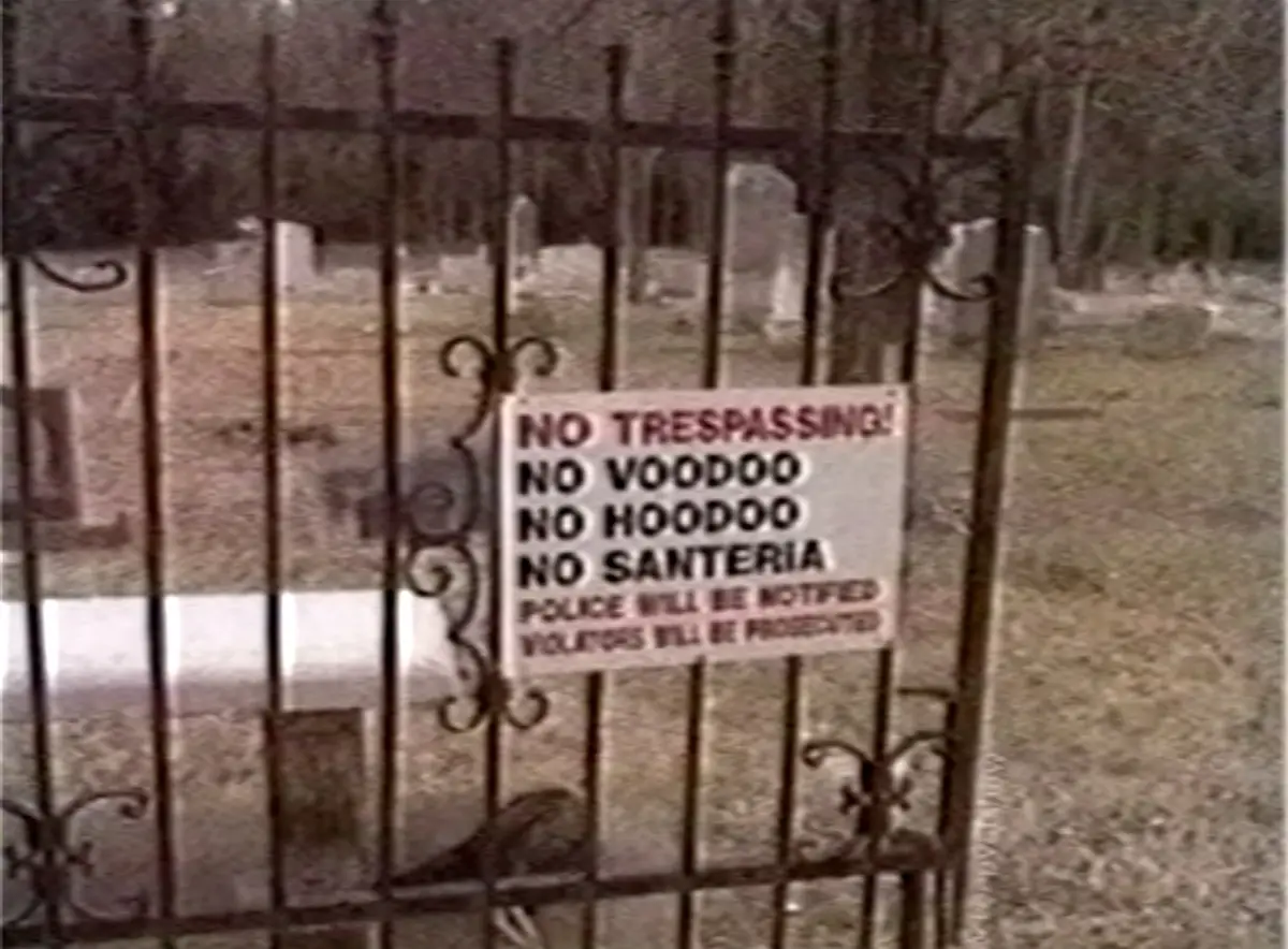A weathered sign attached to a black metal fence in a cemetery lists prohibitions: 'NO TRESPASSING!', 'NO VOODOO', 'NO…