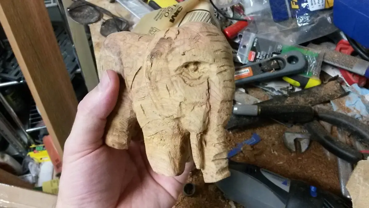 A hand holds a roughly carved wooden elephant head against a cluttered workshop background filled with tools. The overlaid…