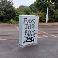 A white-painted concrete block sits in a parking lot, spray-painted with the words 'FUCK THE KING' in bold black letters…